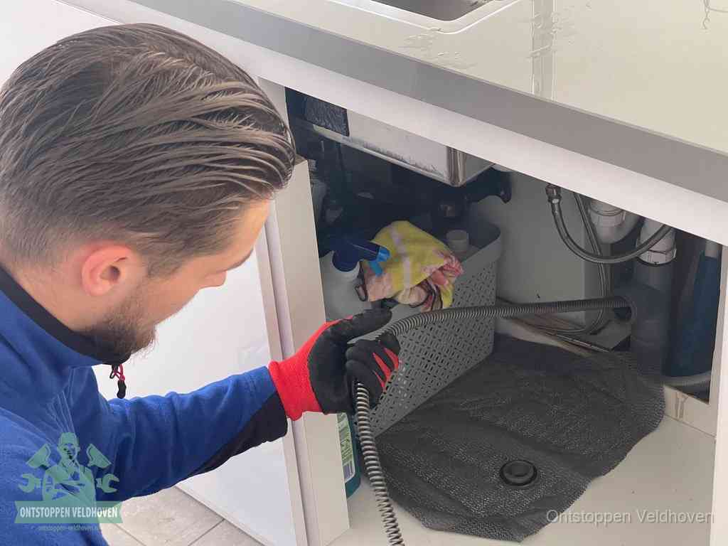 Een monteur voert een flexibele ontstoppingsveer in onder een keukenkastje om een verstopping voorbij de sifon te bereiken in Veldhoven