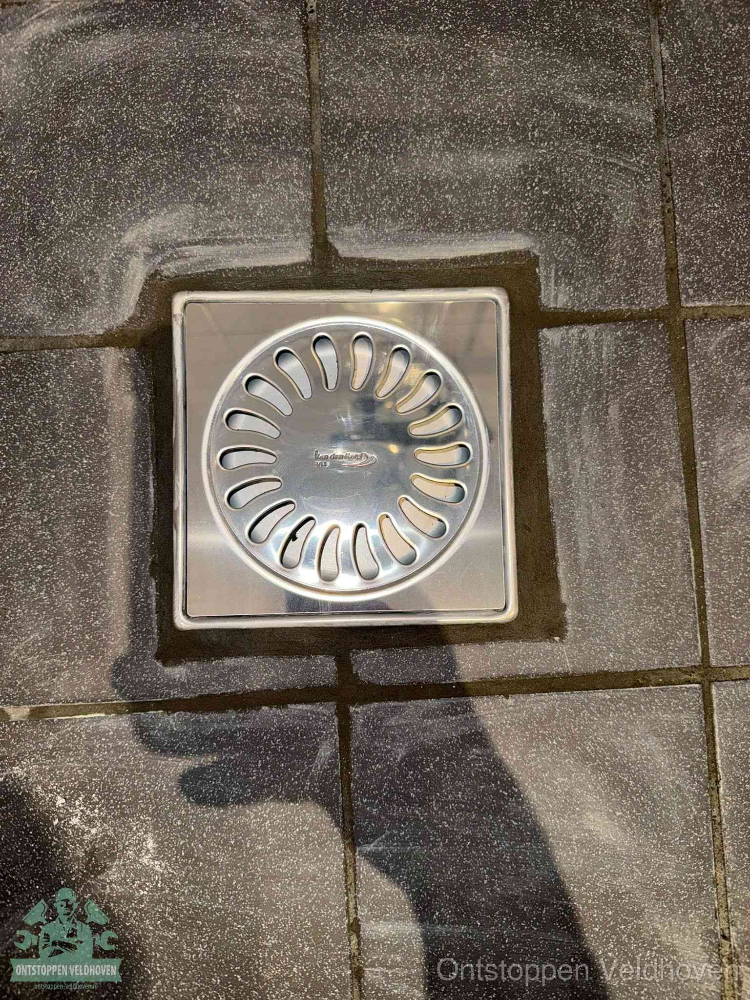 Een schoon vierkant roestvrijstalen vloerputje met sleufrooster in donkere vloertegels, periodiek onderhoud voorkomt een afvoerverstopping in Veldhoven