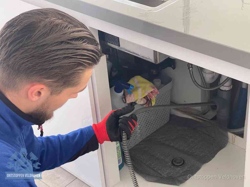 Een monteur voert een flexibele ontstoppingsveer in onder het keukenkastje, langs de sifon, om de gootsteen te ontstoppen in Veldhoven