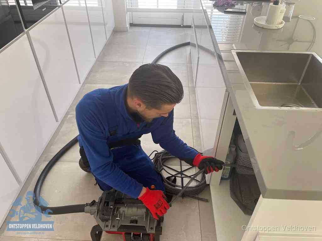 Een monteur ontstopt de keukenafvoer onder de spoelbak met een elektrische veermachine in Veldhoven