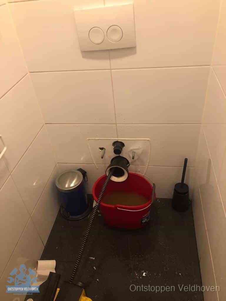 Een ontstopte wand-wc met verwijderde toiletpot waar een ontstoppingsveer in de wanduitgang verdwijnt, het achterhalen waar de verstopping zit in Veldhoven