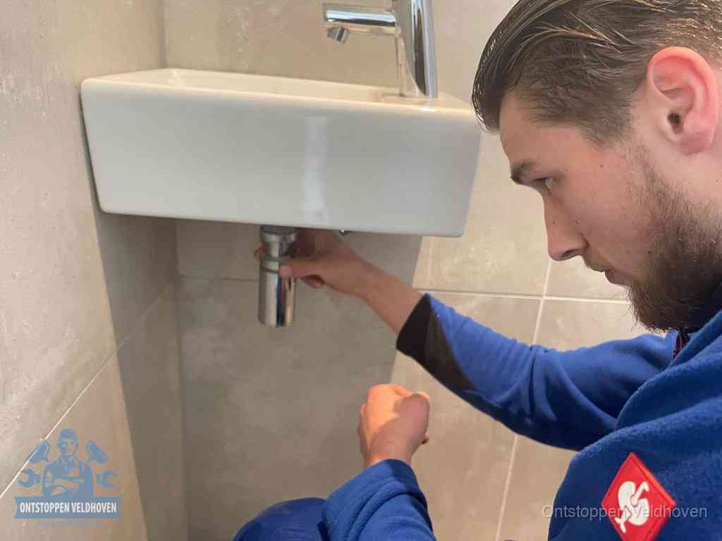 Een monteur draait met de hand de zwanenhalssifon onder een fonteinwasbak in de toiletruimte los om de bron van rioollucht in Veldhoven te vinden