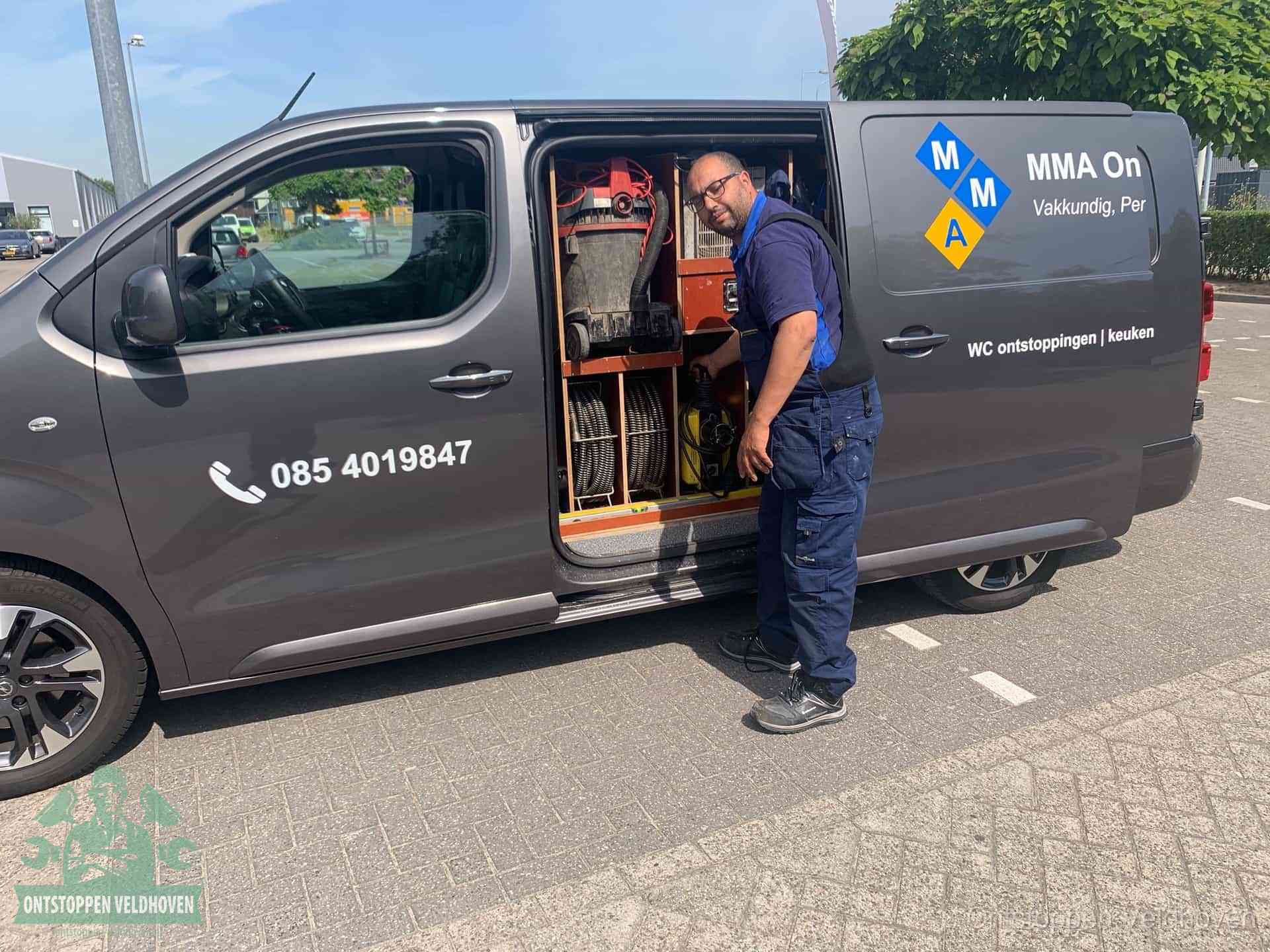 Een monteur bij de open schuifdeur van zijn bedrijfsbus op een parkeerplaats, klaar om de oorzaak van terugkerende afvoerverstoppingen aan te pakken in Veldhoven