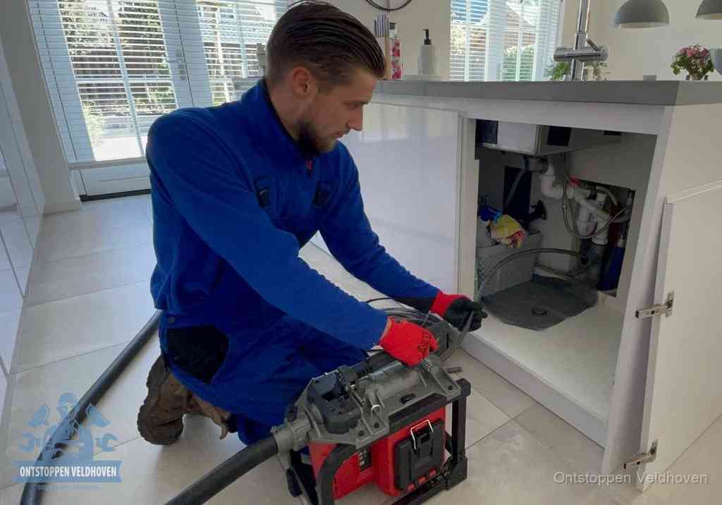 Een monteur voert een professionele elektrische ontstoppingsmachine in de afvoer onder de spoelbak tijdens het gootsteen ontstoppen in Veldhoven